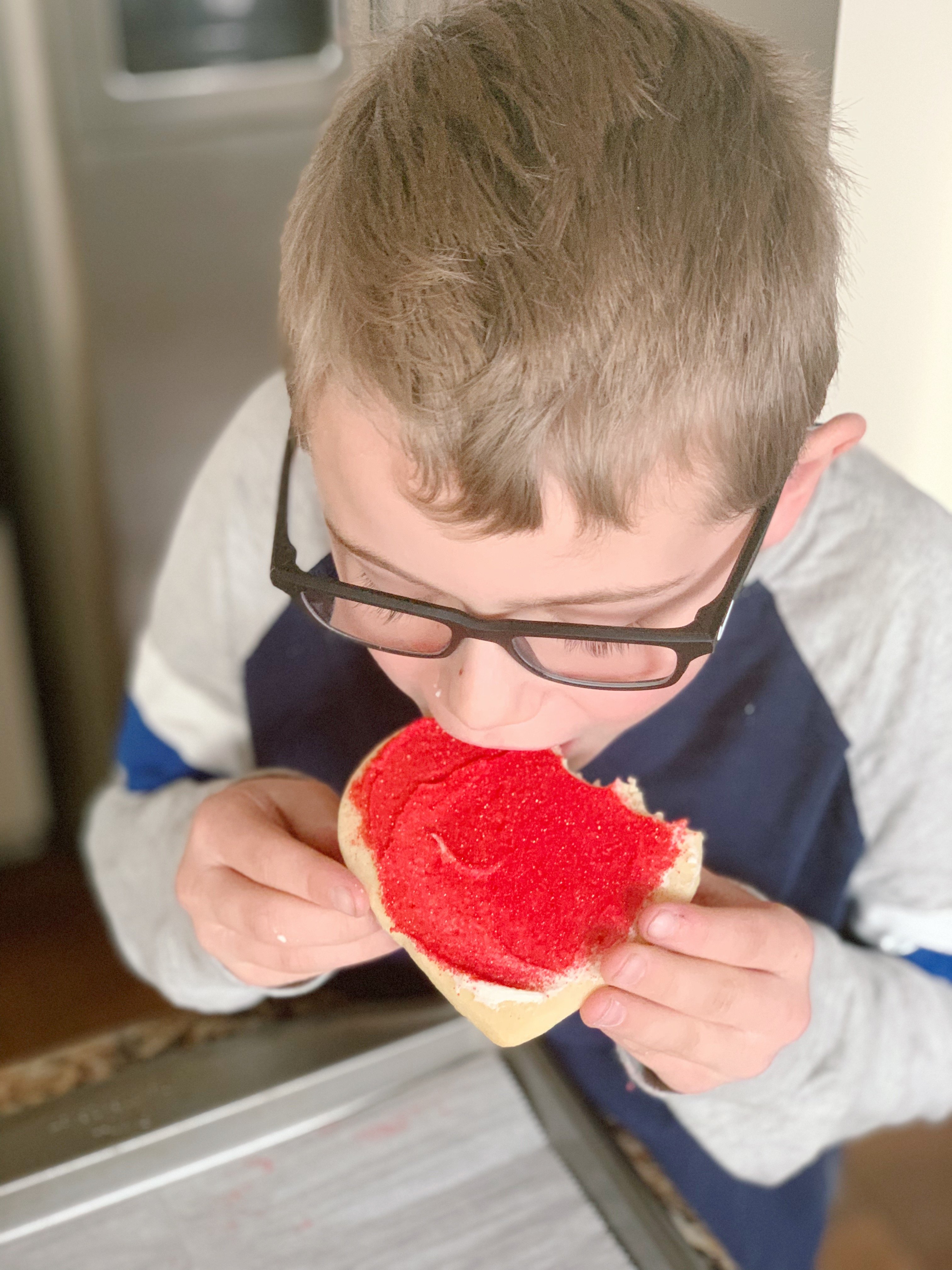 Blonde boy with navy and grey shirt eating a square sugar cookie with red frosting.