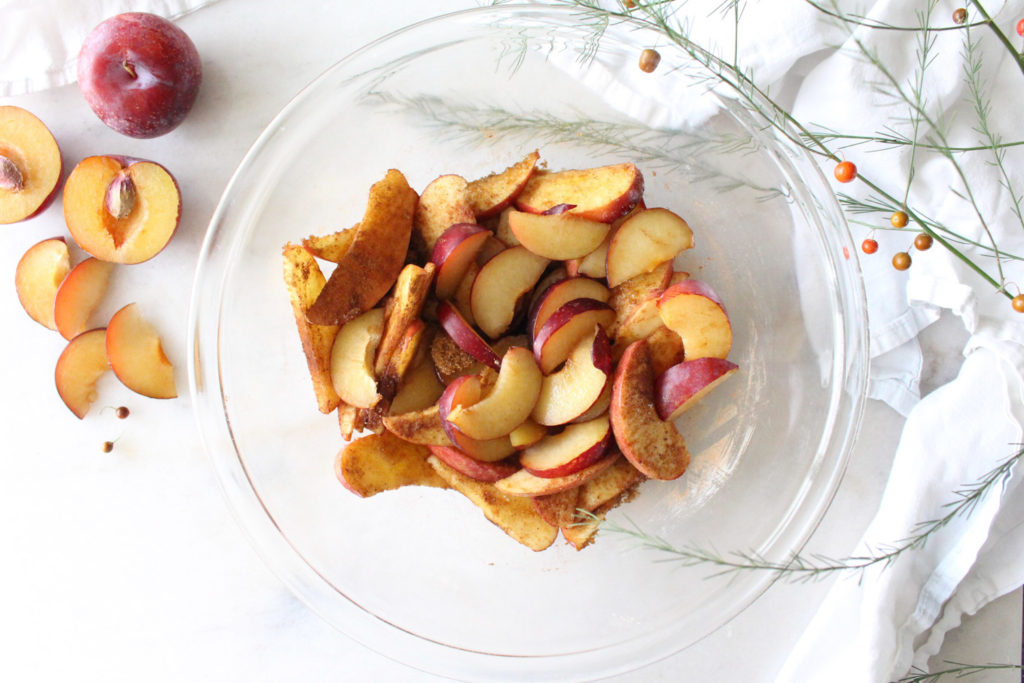 Sliced apples and plums in a glass mixing bowl, combined with brown sugar and cinnamon to fill a galette.