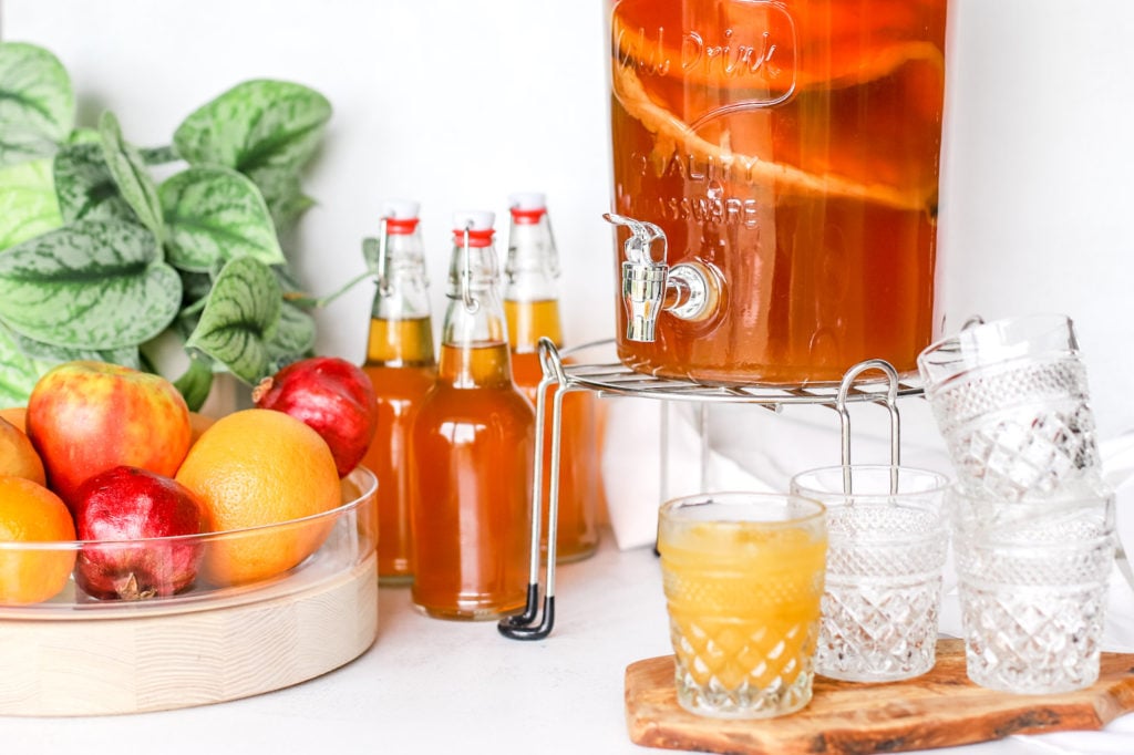 Kombucha fermentation vessel and bottled drinks with a bowl of fruit and beverage dispenser.
