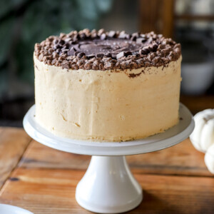 A layered peanut butter cake on a white cake stand, frosted with peanut butter buttercream and topped with a thing layer of fudge and crumbled chocolate chunks.