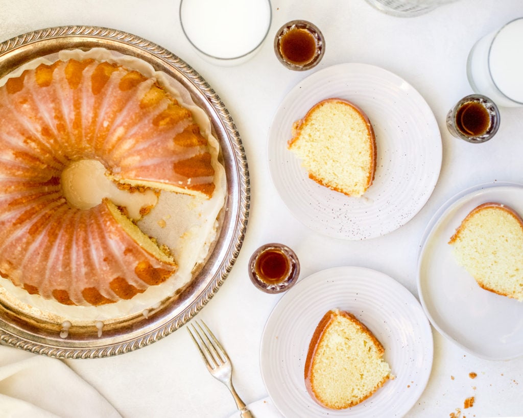 A sliced bundt cake on a silver platter with three slices on plates with shots of bourbon.