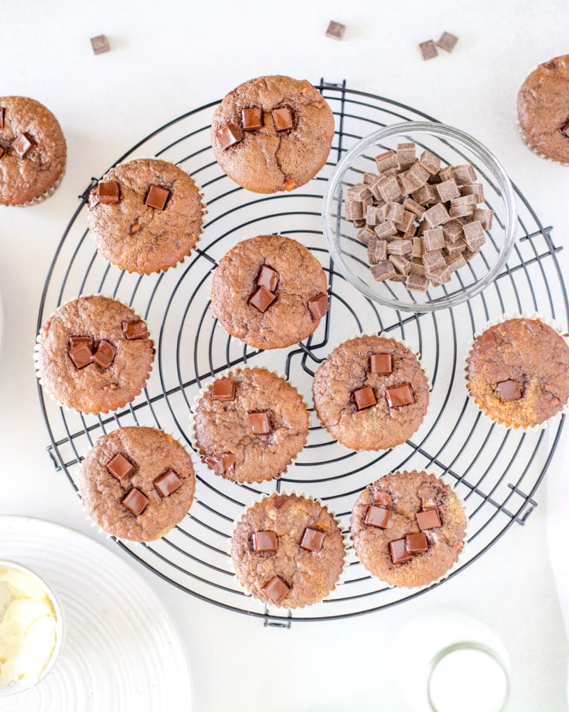 Freshly baked chocolate banana muffins on a round cooling rack with a bowl of chocolate chips.