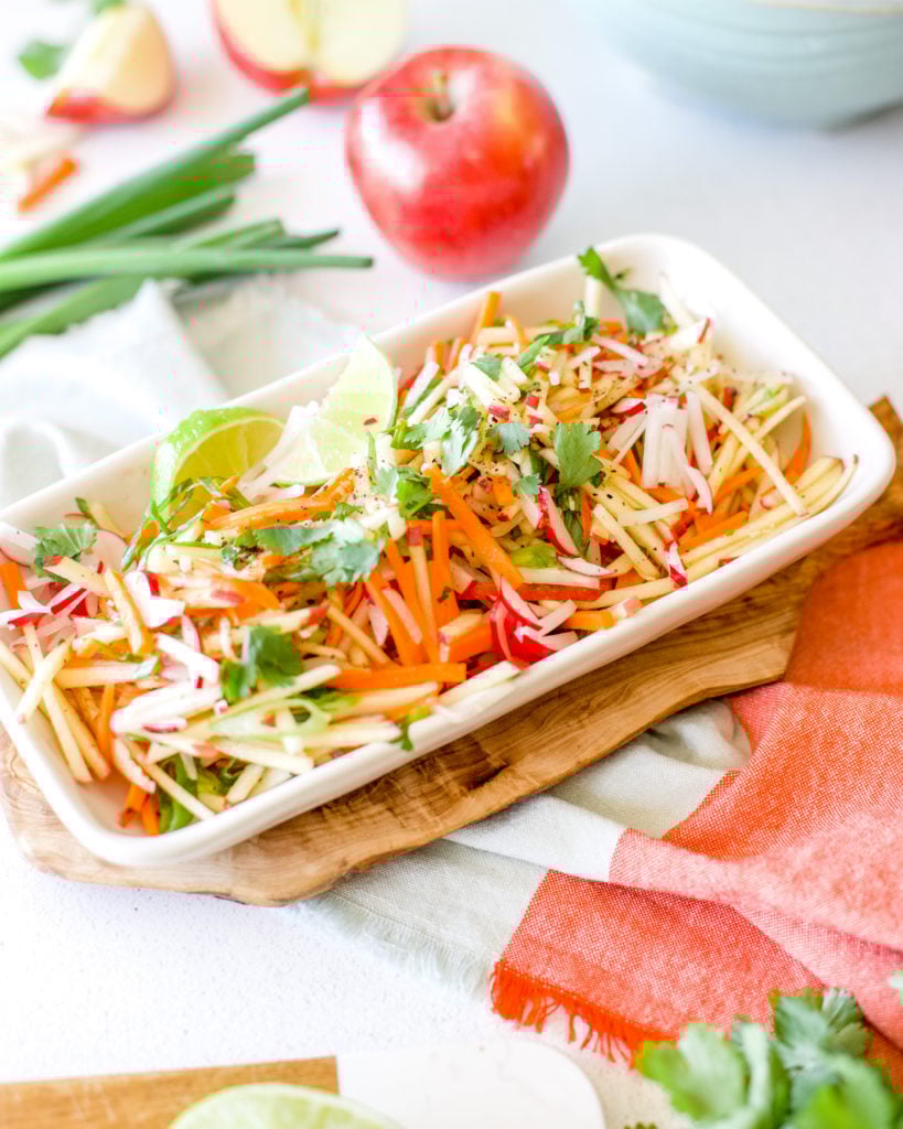 Apple slaw arranged on a rectangular platter with wedges of lime for garnish.