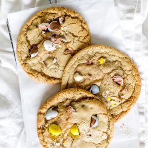 Three Easter cookies with pastel colored mini Cadbury eggs styled on a piece of white parchment paper and a cooling rack.
