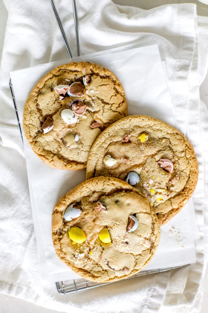 Three Easter cookies with pastel colored mini Cadbury eggs styled on a piece of white parchment paper and a cooling rack.