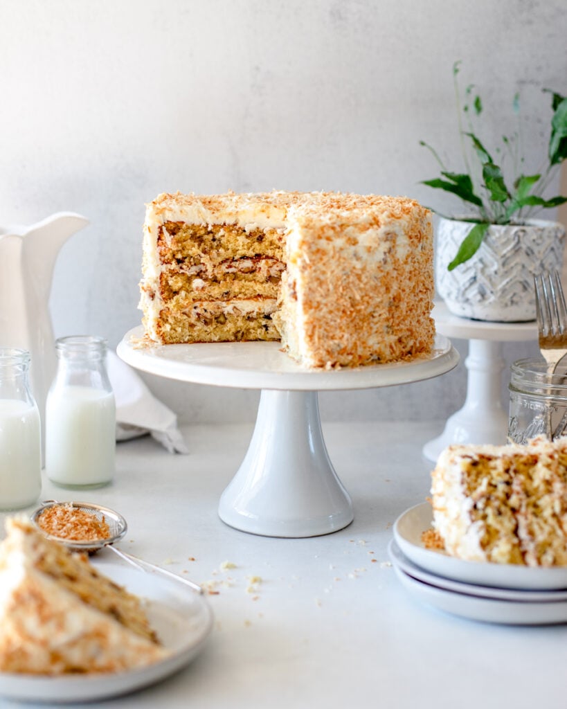A layered Italian Cream Cake on a white cake stand with two sliced pieces and glasses of milk.