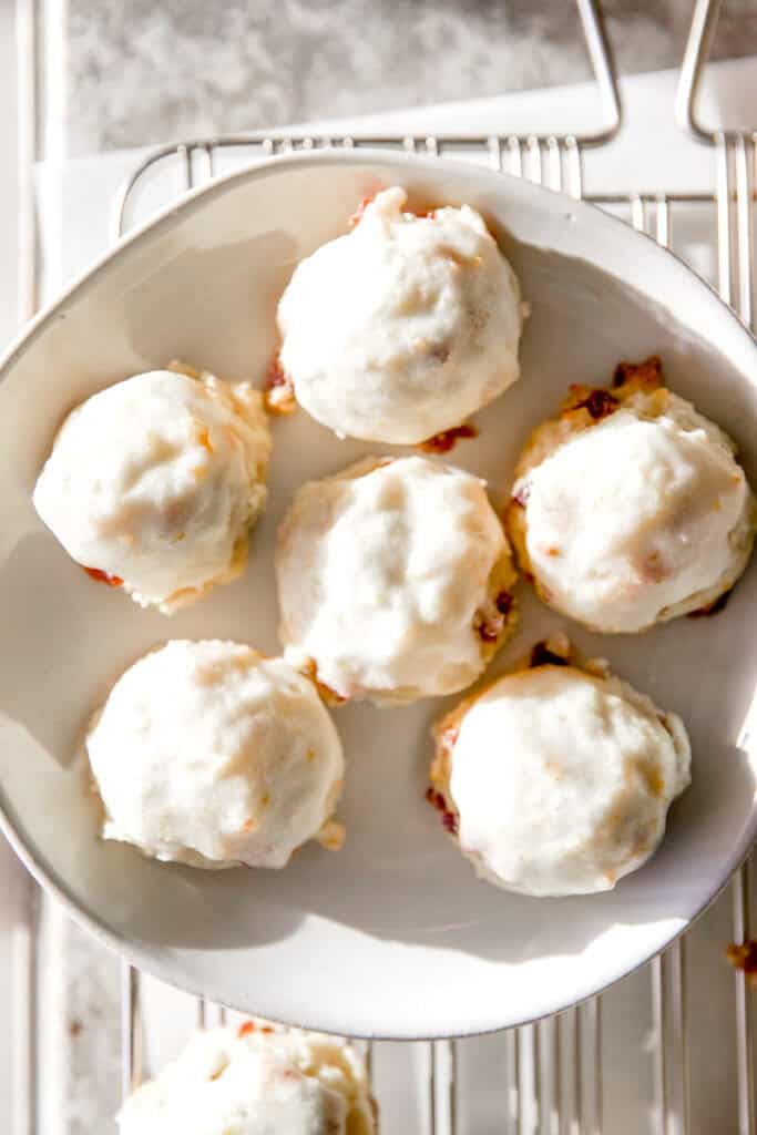 A white stoneware plate with six cranberry orange cookies with orange icing and sprinkles of orange zest and tiny cranberry pieces decorating the cookies.