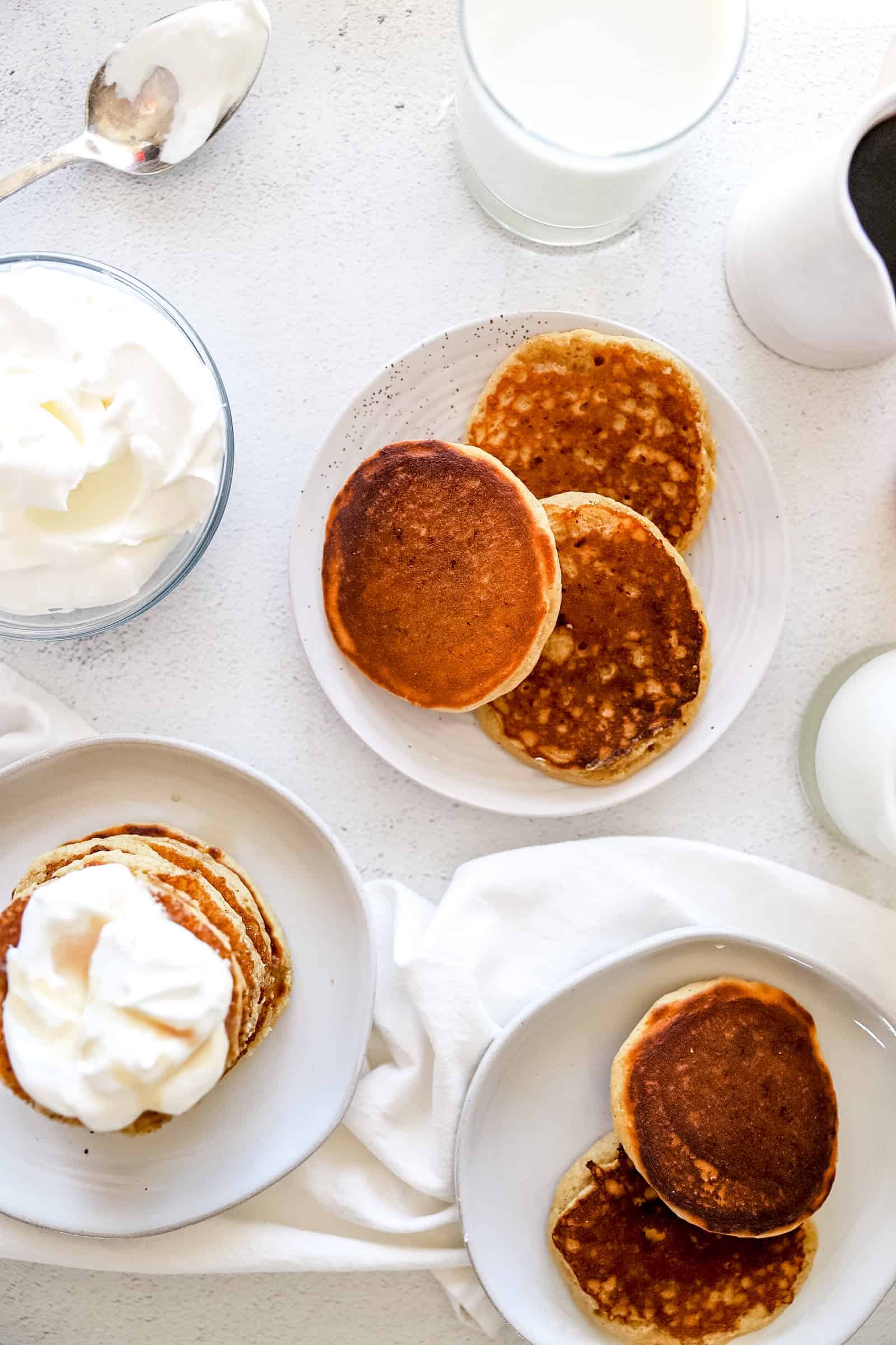 A birds eye view of plates of pancakes with whipped topping garnishes and maple syrup on a white background.