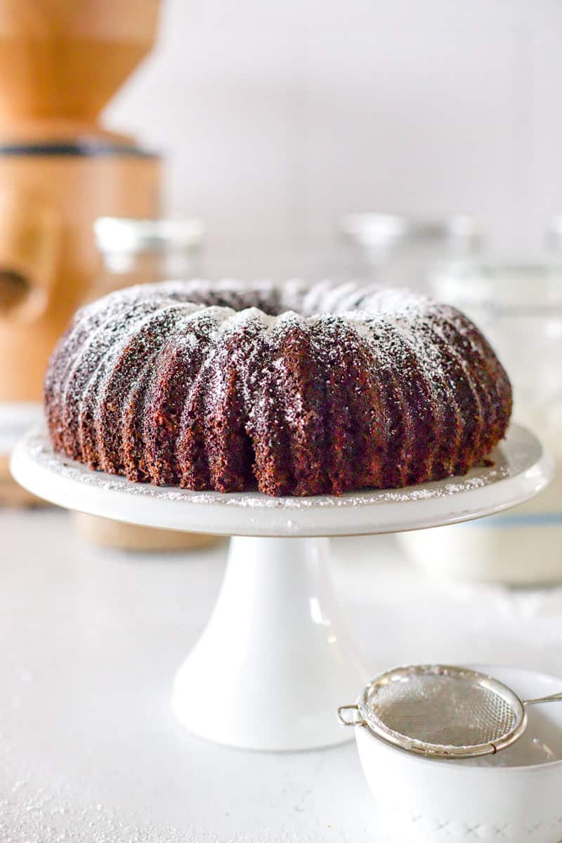 A fudge chocolate sourdough bundt cake on a white cake stand dusted with powdered sugar.