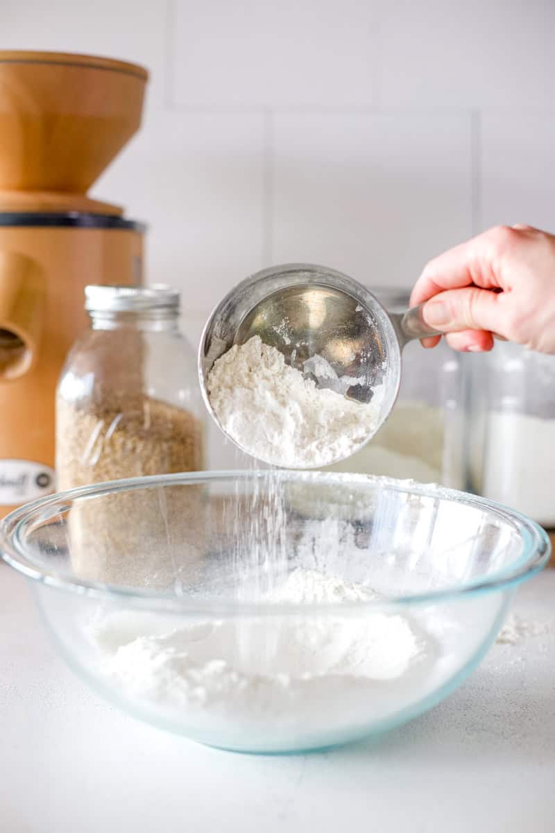 A woman's hand holding a measuring cup full of flour and pouring it into a glass bowl.