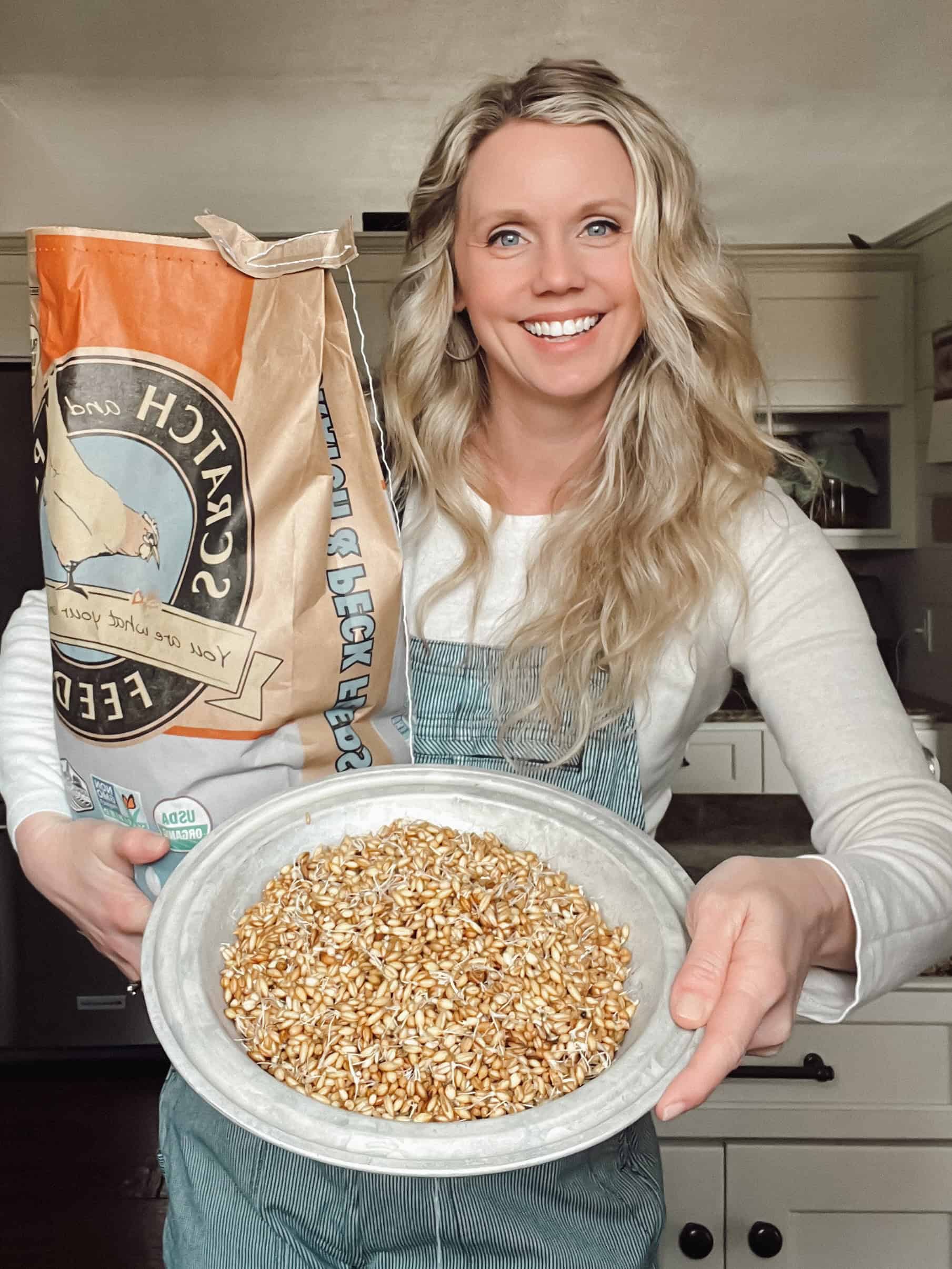 Megan holding a bag of scratch grains in one hand, a galvanized pan of sprouted grains for her chickens in the other hand.