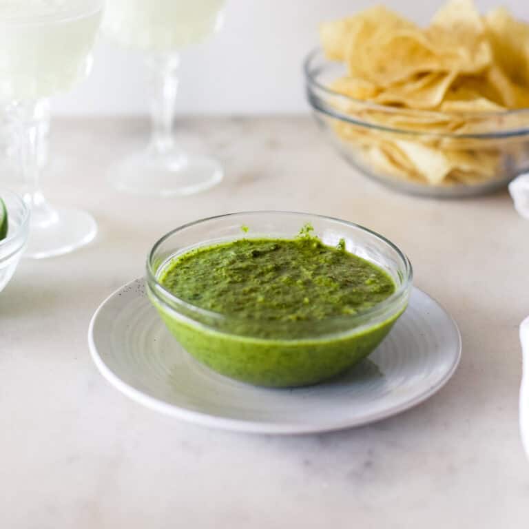 A glass bowl of bright green chimichurri sauce on a white kitchen background with chips and margaritas in the background.