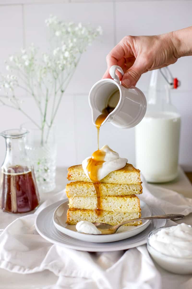 A woman's hand pouring maple syrup over a stack of French toast topped with whipped cream.