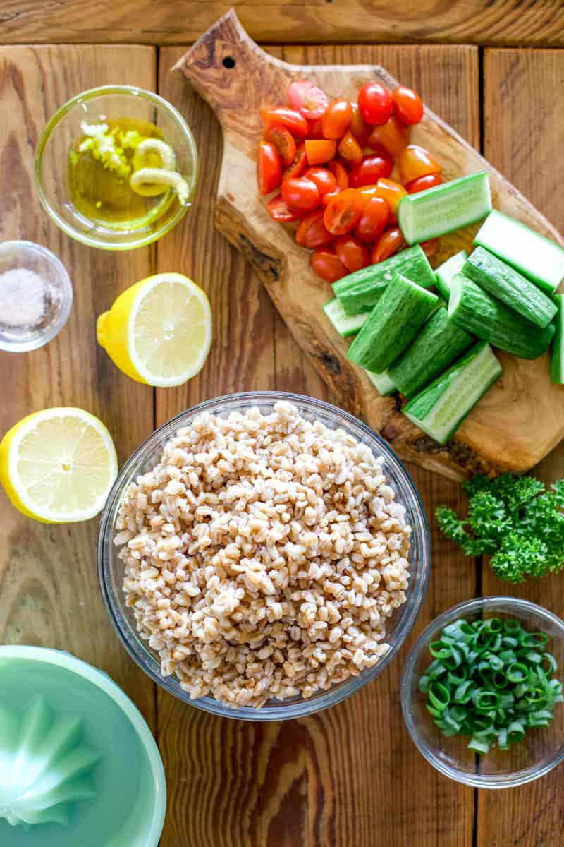 Cooked farro in a bowl with a serving suggestion showing lemon slices, cherry tomatoes, cucumbers, lemon salad dressing and parsley for step four of cooking farro.