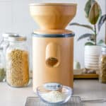 A grain mill grinding wheat with jars of wheat berries in the background.