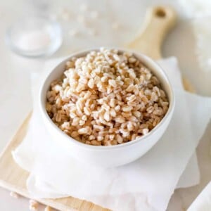 A white bowl of cooked farro on a white background with a serving board and a pinch of salt.