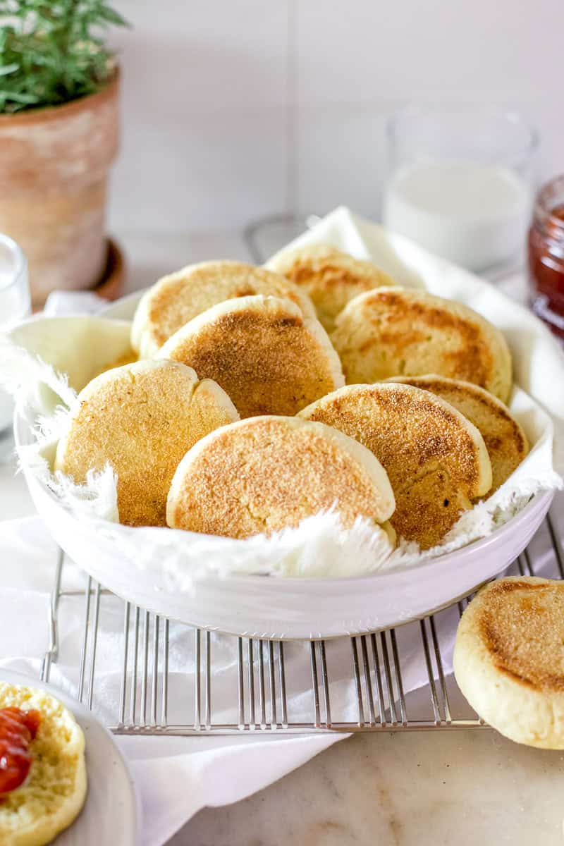 A bowl full of freshly baked sourdough English muffins in a white bowl with a white tea towel tucked underneath them.