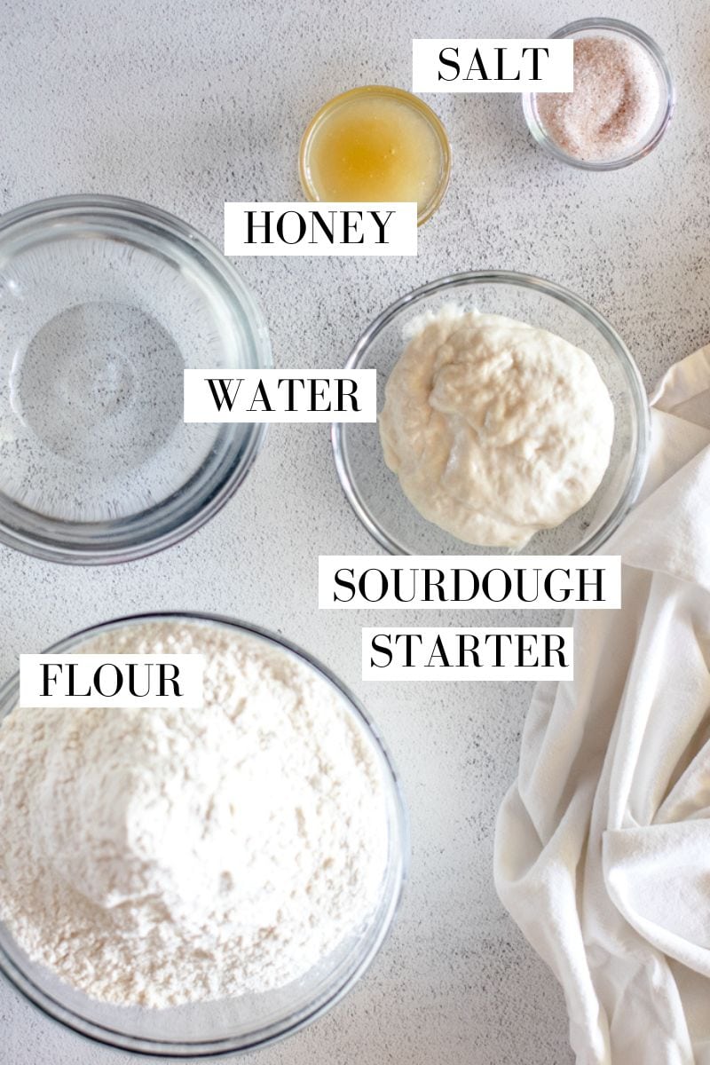 A flat lay view of the ingredients for a sourdough batard including honey, salt, water, sourdough starter and flour in glass prep bowls.