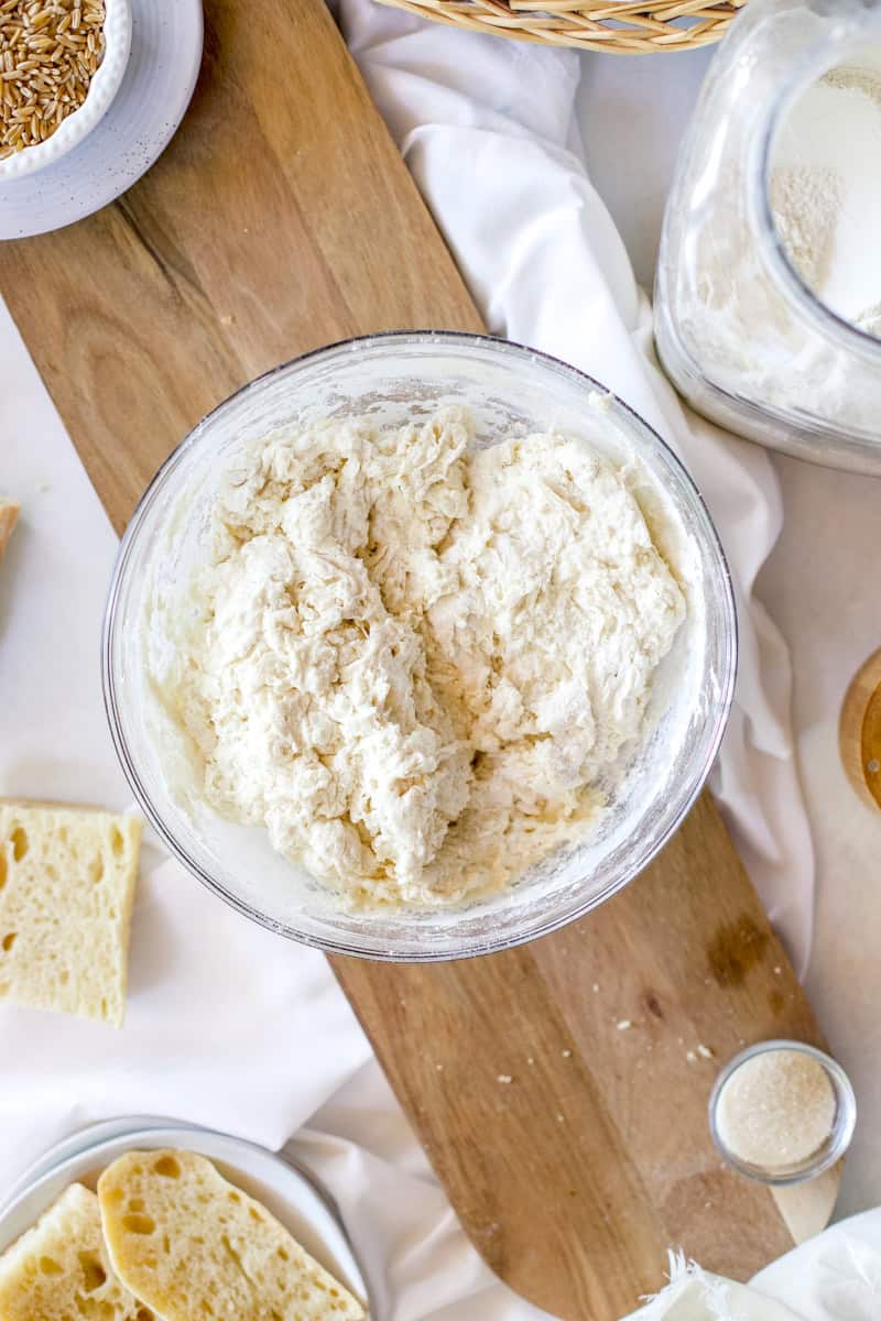 A photo of step 2 of instructions for making sourdough baguettes is adding the flour, sugar, and salt to the bowl of water and sourdough starter.