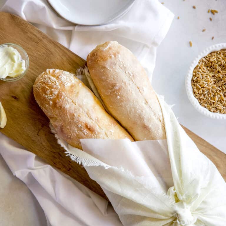 Two loaves of 5 ingredient sourdough baguettes wrapped in linens and parchment paper on a wooden serving board.