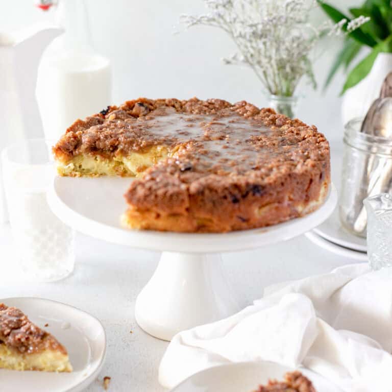 A rhubarb sour cream cake on a white pedestal with glasses of milk, serving plates and vintage forks.