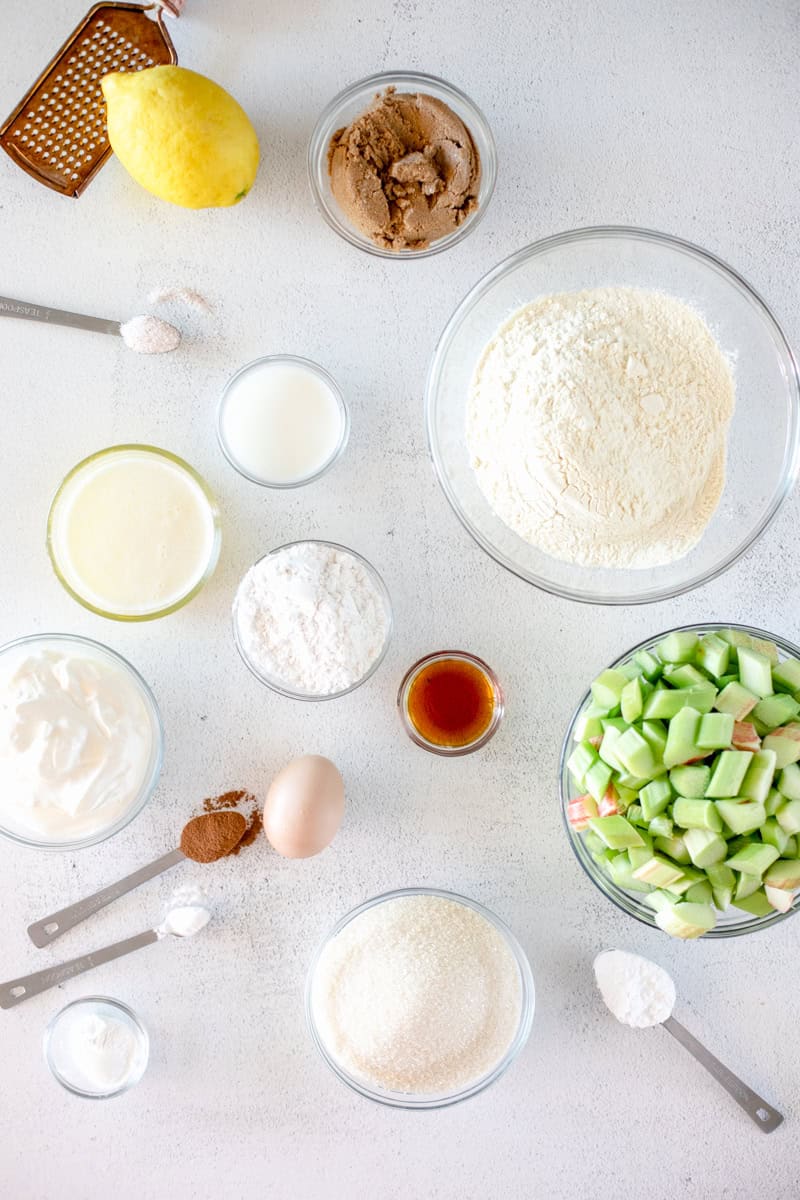 An image showing the ingredients for a rhubarb sour cream cake including flour, sugar, butter, salt, rhubarb and many others in glass prep bowls.