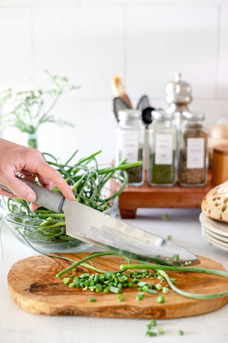 A hand cutting garlic scapes into small pieces on a wooden cutting board.