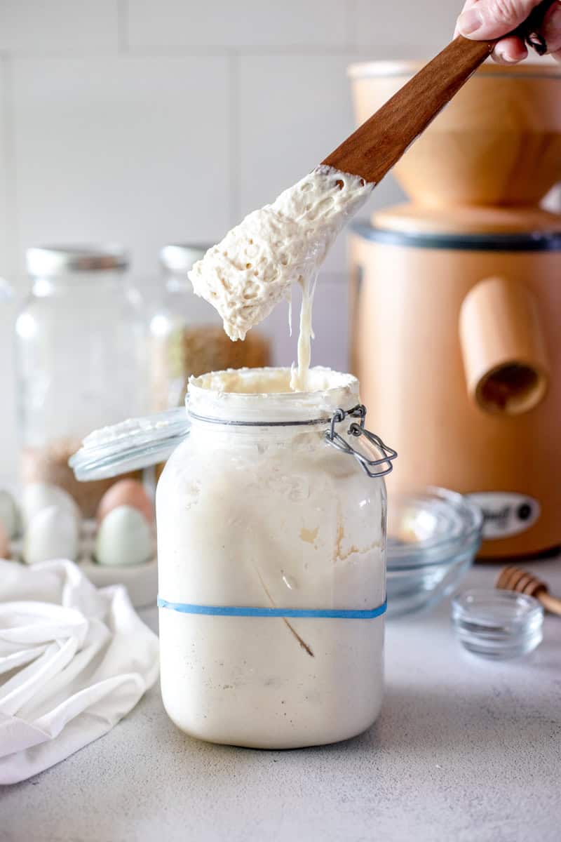Sourdough starter in a large glass jar with a flip-top being stirred by a wooden sourdough spoon with a Mockmill grain mill in the background.