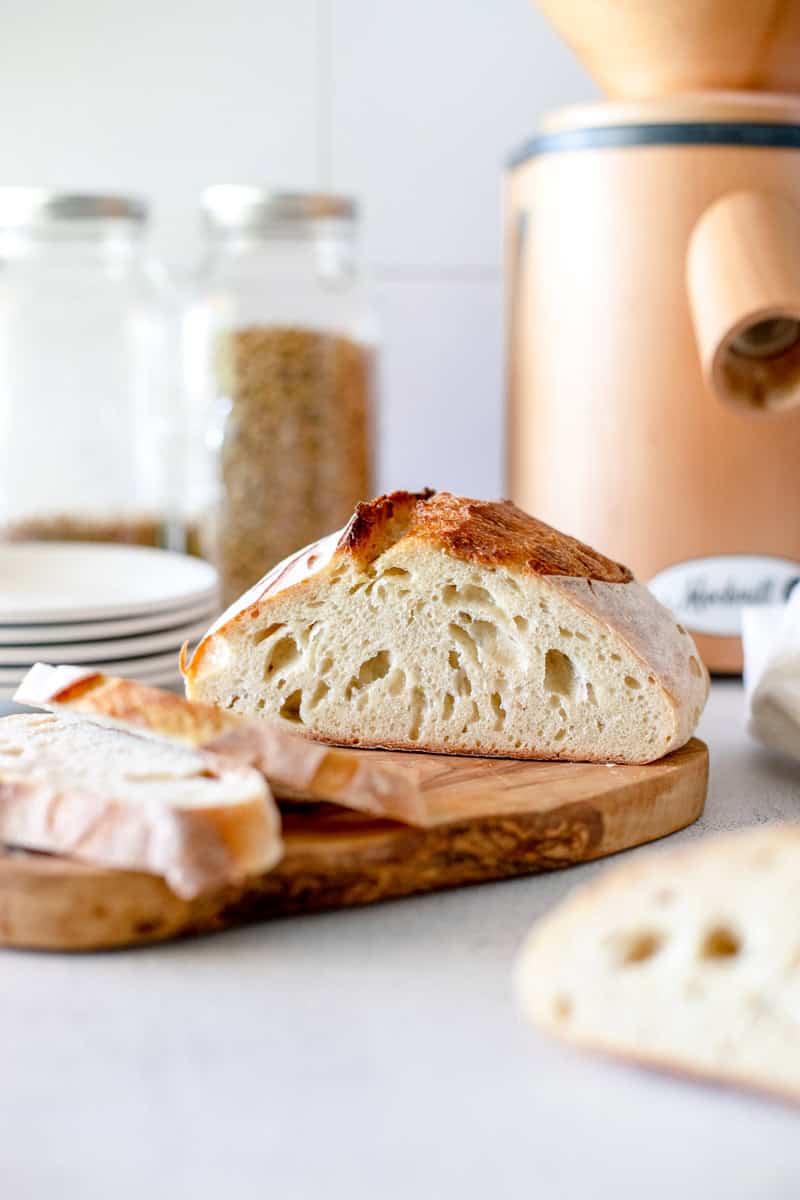 A fresh sourdough batard sliced on a wooden cutting board with a jar of wheat and grain mill in the background.