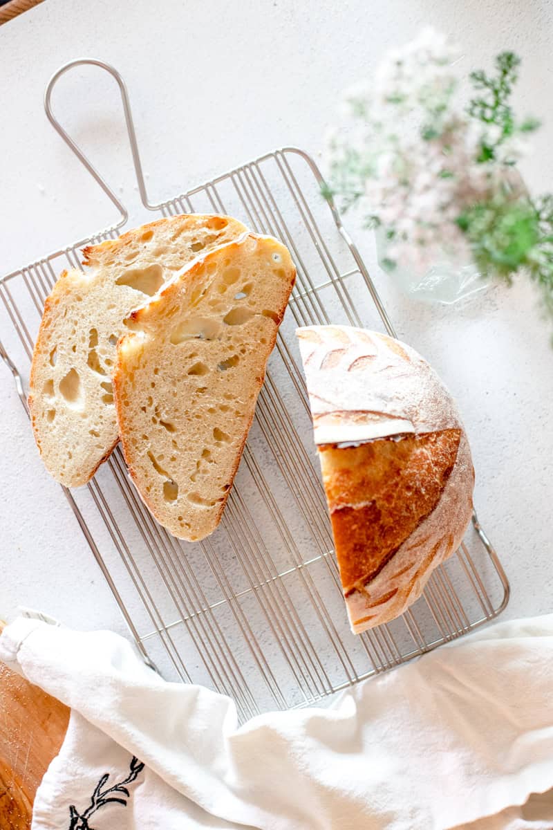A sliced sourdough batard on a wire cooling rack with some tiny white flowers in a vase.