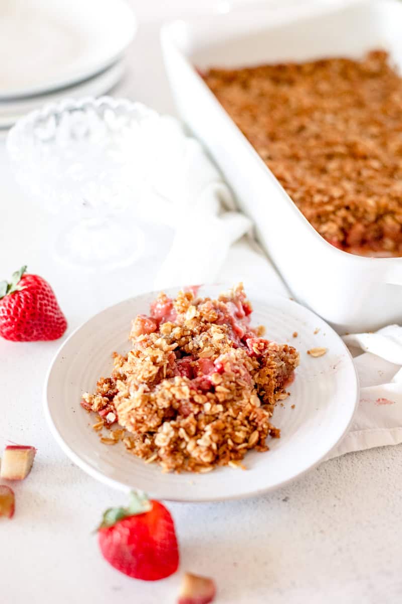 A slice of freshly baked rhubarb strawberry crisp on a white plate with the baking dish behind it.