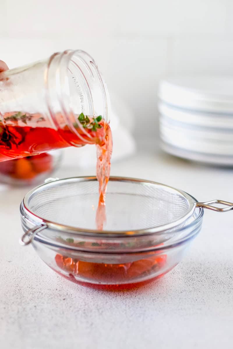 Step three of making strawberry infused vinegar is straining the mason jar full of strawberry tops into a mesh sieve into a bowl.