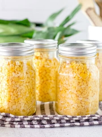 Pints of pressure canned corn in jars with ears of corn in the background.