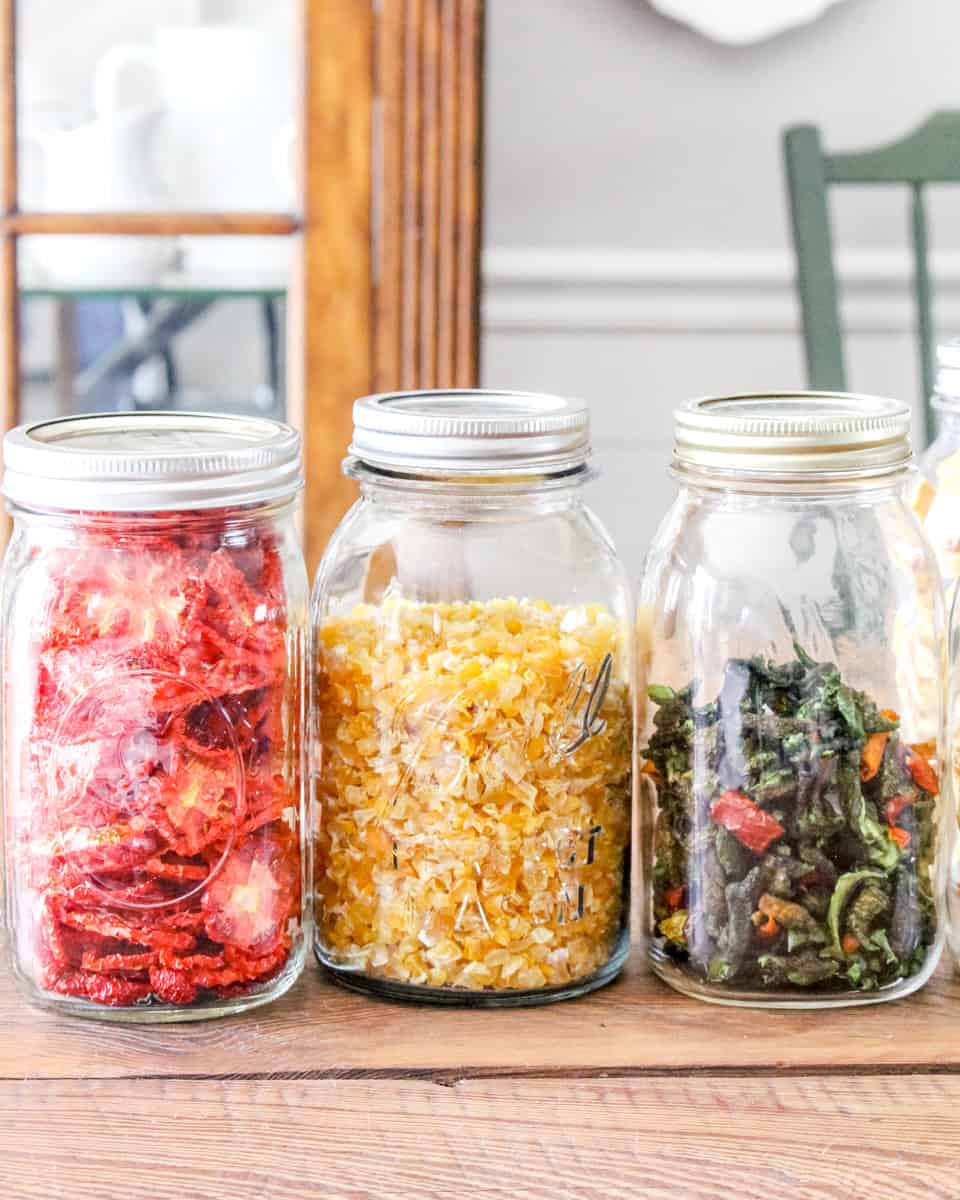 Dehydrated corn, tomatoes and peppers in mason jars on a rustic wooden kitchen table.