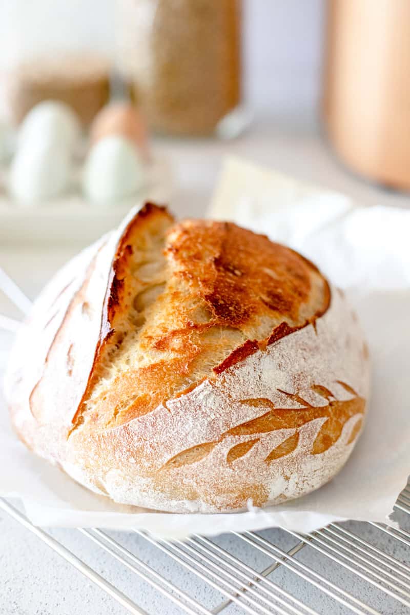 A loaf of sourdough bread that is scored with a leaf pattern on a white parchment and metal wire rack.