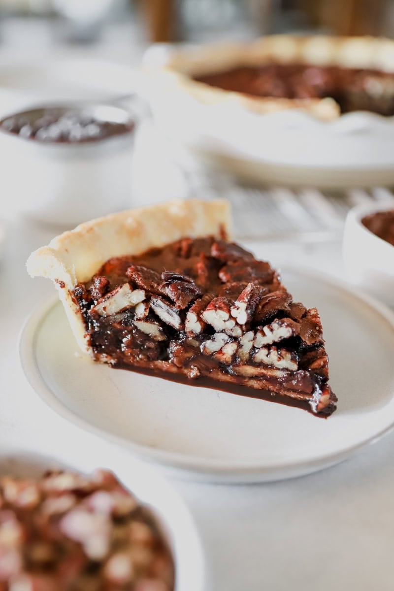 A slice of chocolate pecan pie on a white plate showing the detail of the nuts and fudgy filling.