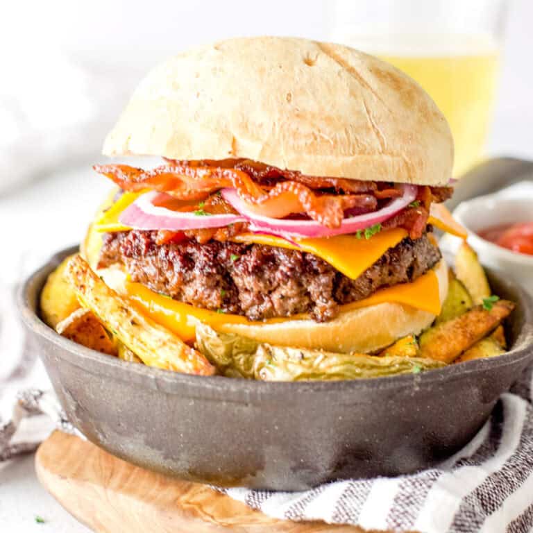 a juicy venison burger served with cheddar cheese and oven fries with slices of thick cut bacon. It's being served up in a mini cast iron skillet over a bed of fresh oven fries with a frosty glass of beer.
