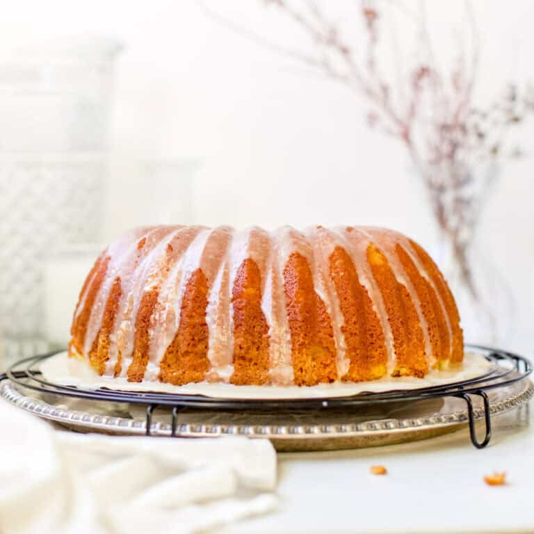 a beautiful glazed bundt cake on a cooling rack with dried flowers in a glass vase blurred in the background.
