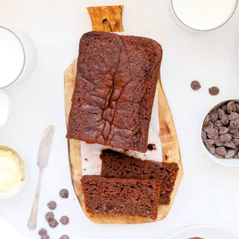 A freshly baked loaf of chocolate sourdough banana bread on a wood cutting board with chocolate chips, creamy butter and a glass of milk.