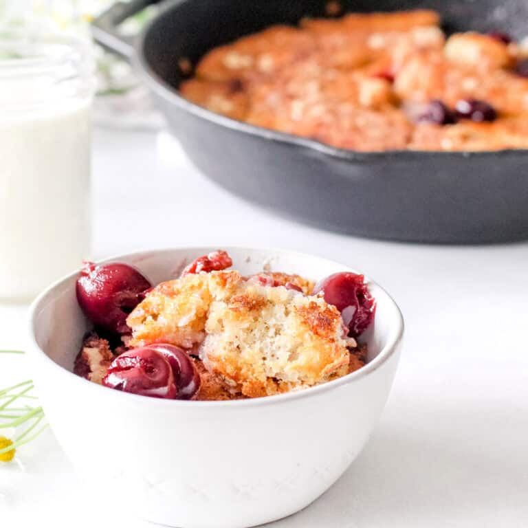 A quick cobbler in a white dish with dark cherries and a cast iron skillet in the background.