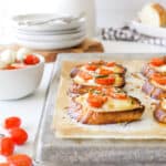 Hot tomato and mozzarella crostinis fresh from the oven on an aluminum baking sheet with tomatoes and basil for garnish.