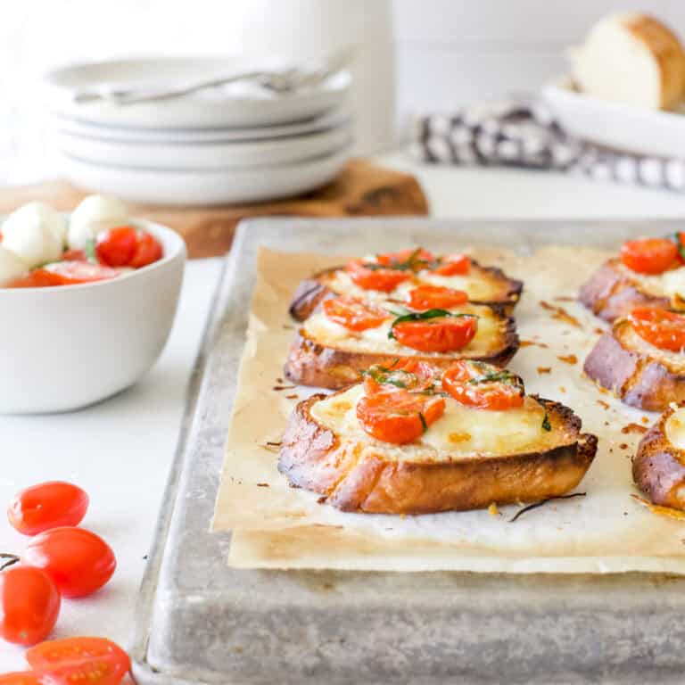 Hot tomato and mozzarella crostinis fresh from the oven on an aluminum baking sheet with tomatoes and basil for garnish.