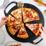 Hands reaching for slices of Italian Grinder Pizza on a Victorria cookware cast iron pizza pan.