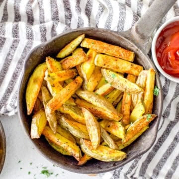A cast iron skillet full of freshly baked easy oven fries on a striped tea towel served with ketchup.