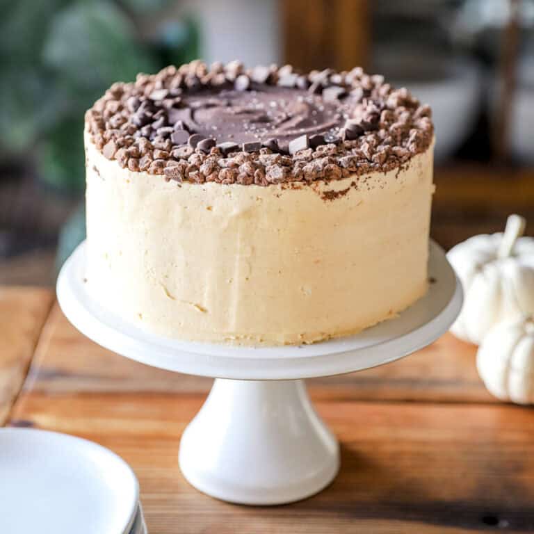 A beautiful peanut butter cake on a pedestal cake plate garnished with chocolate chunks.