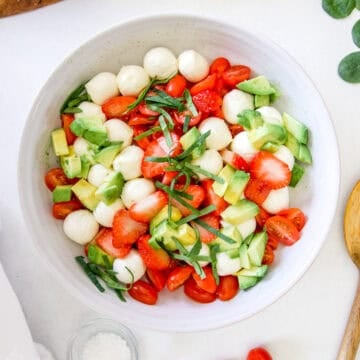 A white stoneware bowl filled with caprese salad made with strawberries, mozzarella, Avocado, basil and tomatoes.