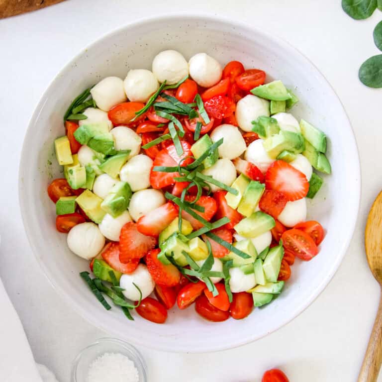 A white stoneware bowl filled with caprese salad made with strawberries, mozzarella, Avocado, basil and tomatoes.