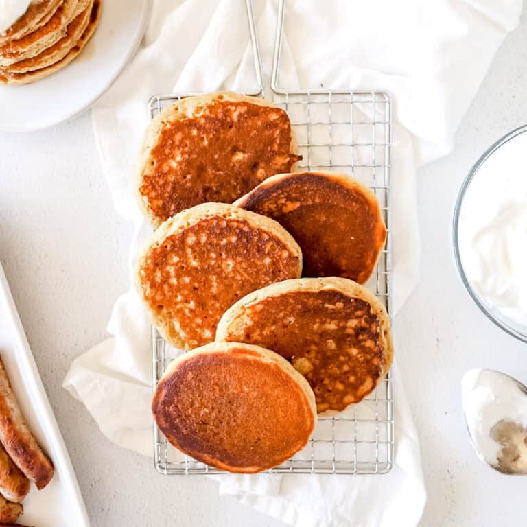 A birds eye view of a wire rack of pancakes with whipped topping garnishes and maple syrup on a white background.