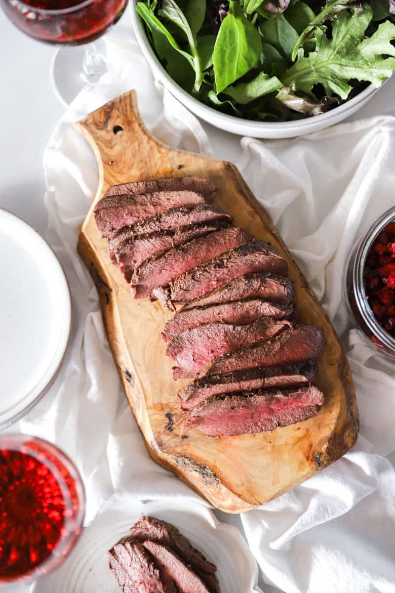 A sliced juicy venison steak on a cutting board with salad and red wine.
