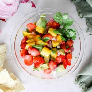 A top view of fresh mango salad in a glass bowl garnished with cilantro, tortilla chips and red onion.